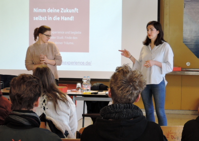 Schulbesuch in Berlin: Zwei Mentorinnen reden vor einer Schulklasse, im Hintergrund ist auf einer Folie der Satz zu lesen: Nimm deine Zukunft selbst in die Hand! (Foto: ArbeiterKind.de Berlin)